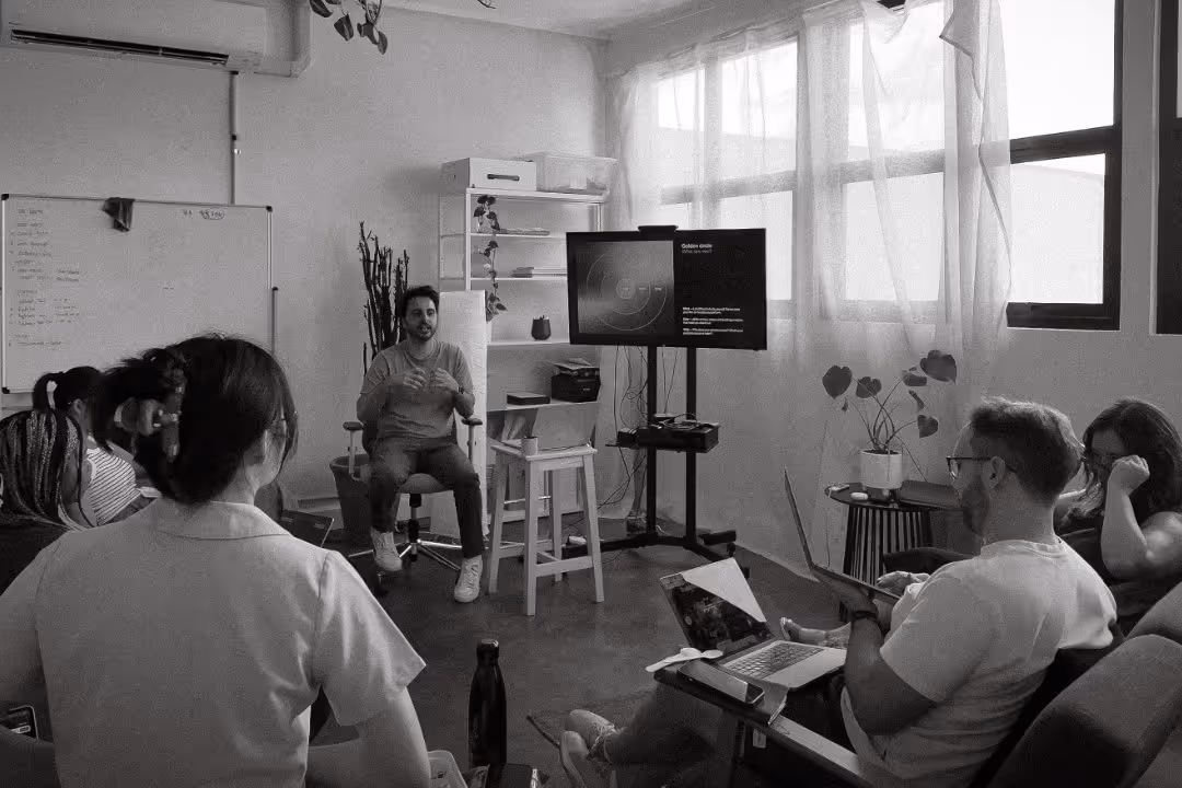 A man seated on a chair leads a workshop with five attendees in a bright room with large windows and a TV display.