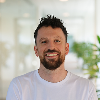 Smiling man with curly hair and beard wearing a white shirt in a bright indoor setting.