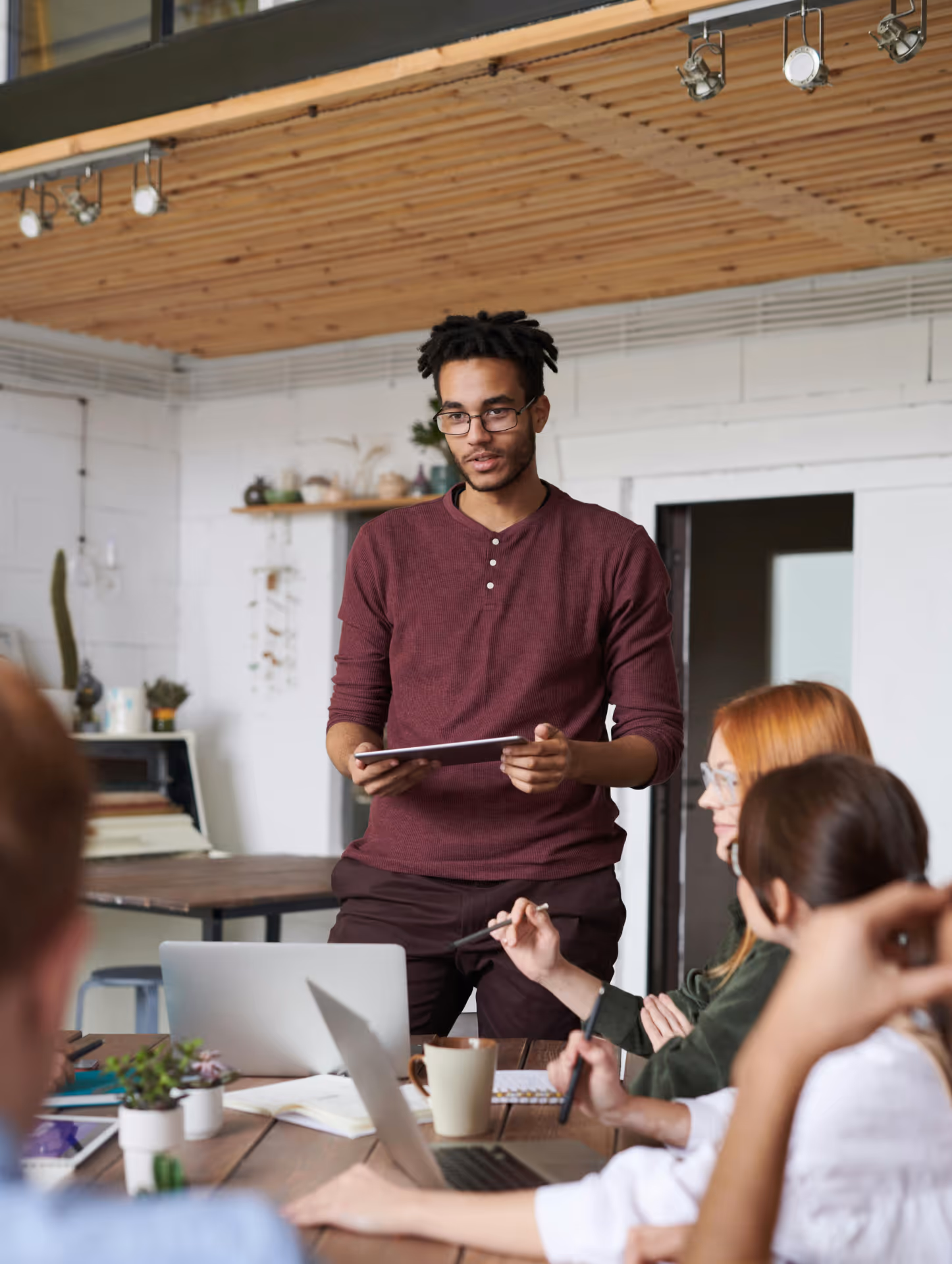 Young man in glasses and maroon shirt holding a tablet presenting to a group of colleagues seated at a table with laptops and notebooks in a modern office.