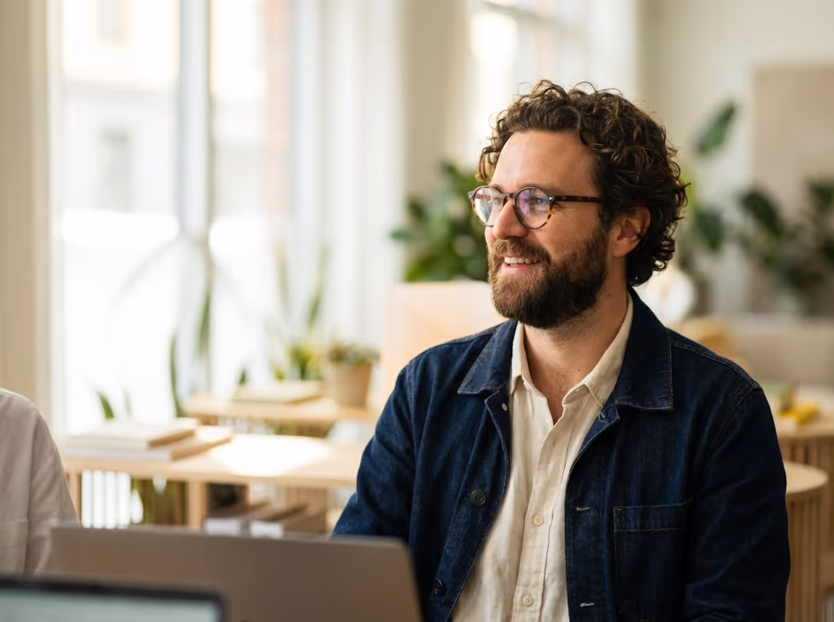 Bearded man with glasses wearing a dark jacket and light shirt, sitting indoors and smiling.