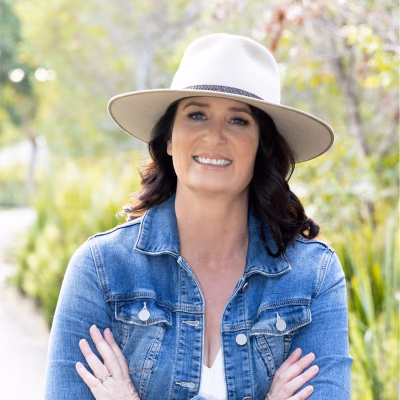 Smiling woman in a white wide-brimmed hat and blue denim jacket with arms crossed, standing outdoors with greenery in the background.