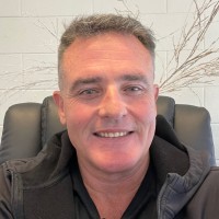 Smiling man with short dark hair sitting in a black chair against a white brick wall with decorative branches.