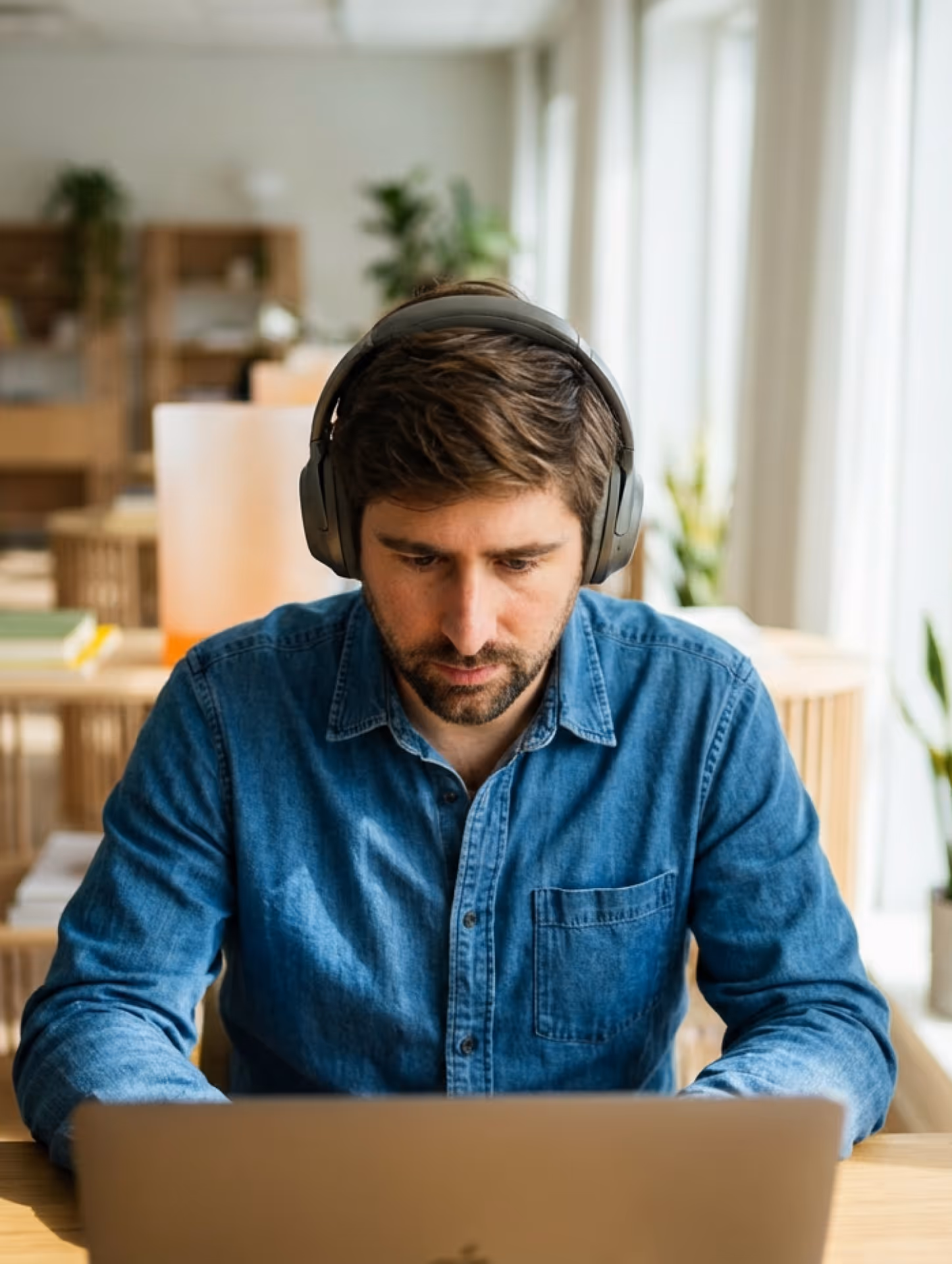 Man wearing headphones and a blue denim shirt working on a laptop in a bright room with plants.