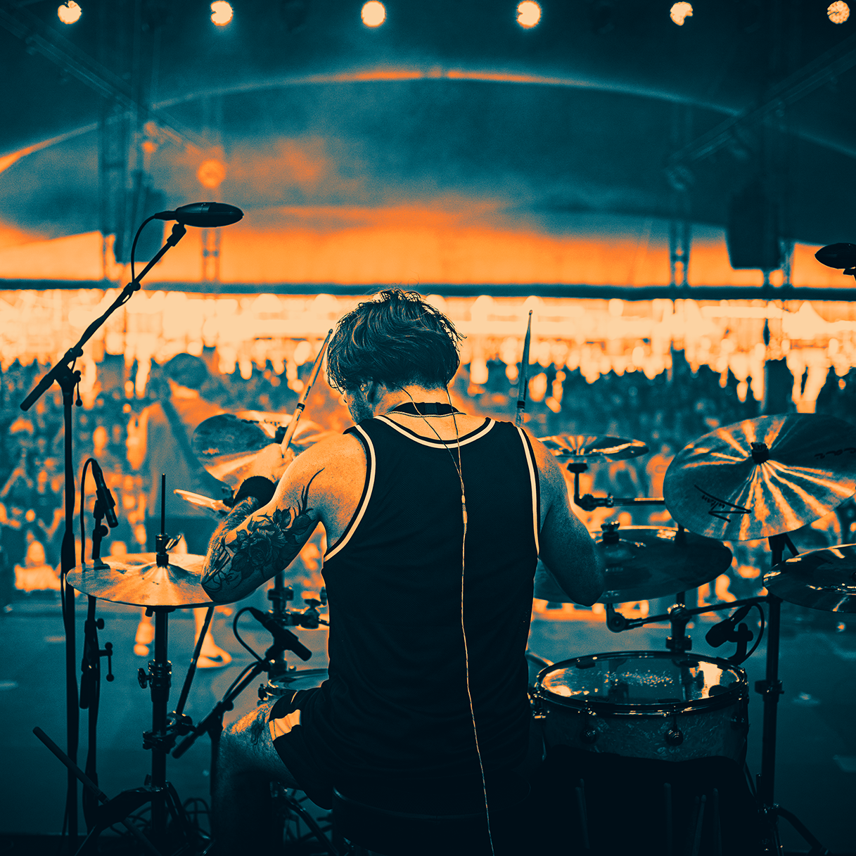Drummer playing on a drum set on stage with hair flying, illuminated by bright orange backlighting and a crowd in the background.