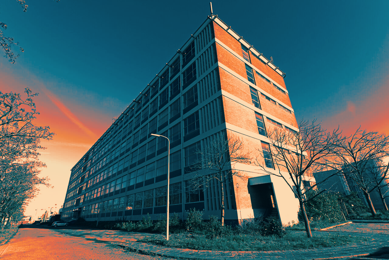 Wide-angle view of Plan-B in Eindhoven, a large multi-story brick and glass building with leafless trees and a street lamp along the sidewalk under a clear sky at sunset.