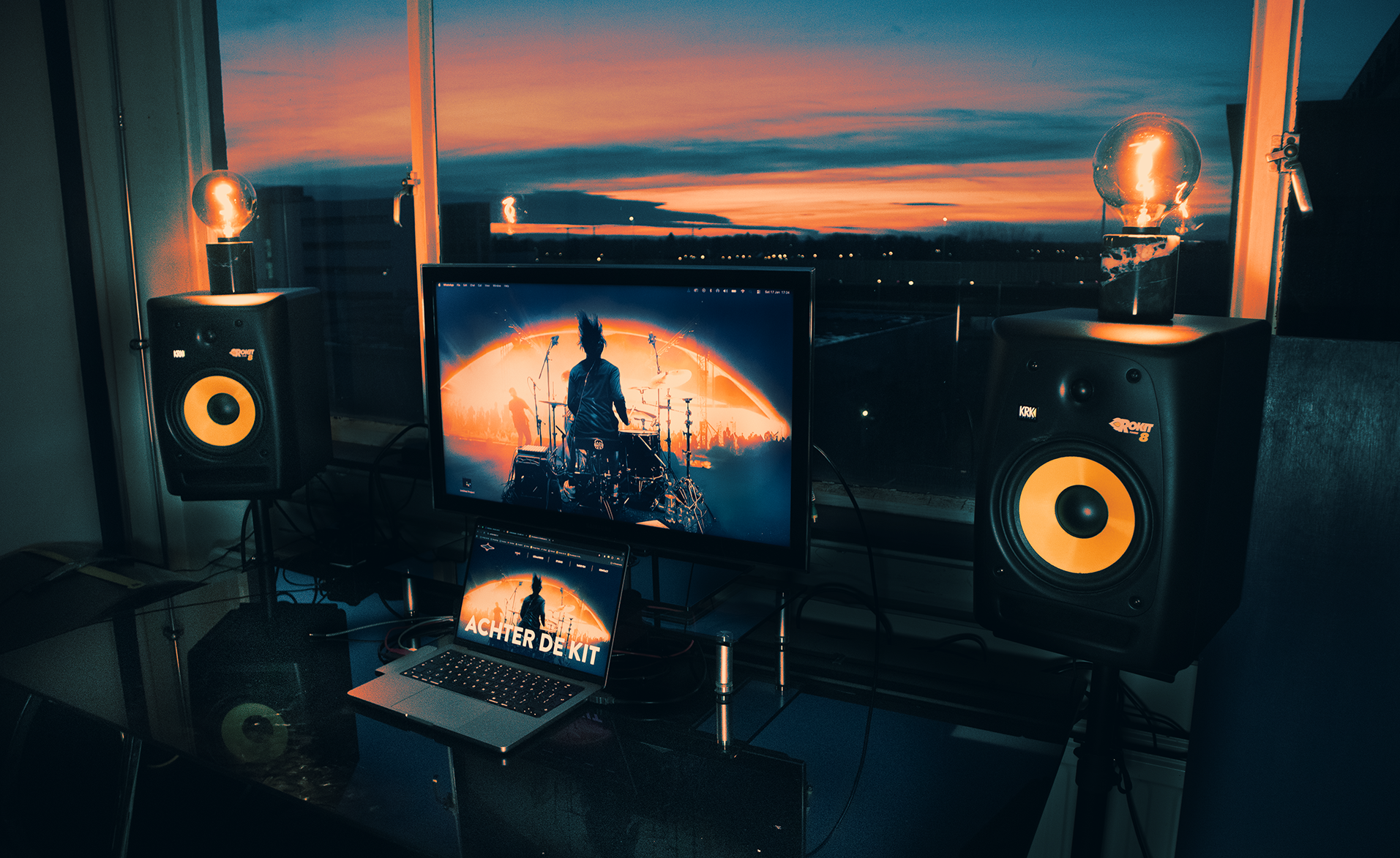 Modern desk setup with a laptop and large monitor displaying a drummer on stage, flanked by two studio speakers topped with vintage filament bulbs, in front of window blinds.