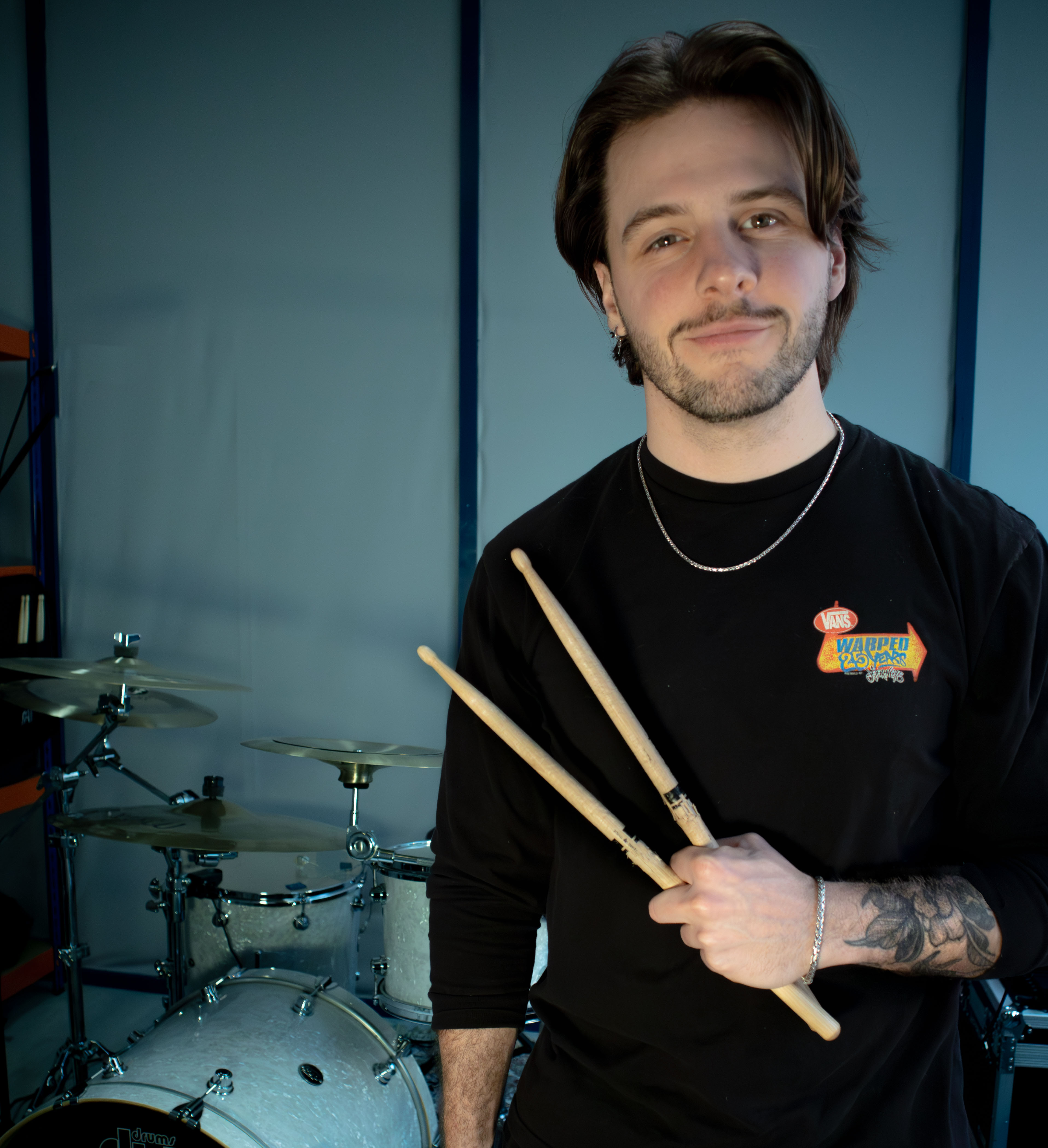 Modern desk setup with a laptop and large monitor displaying a drummer on stage, flanked by two studio speakers topped with vintage filament bulbs, in front of window blinds.