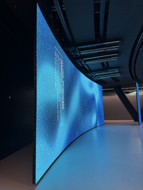 Large curved digital display screen with blue textured background and white text in a modern exhibition space.