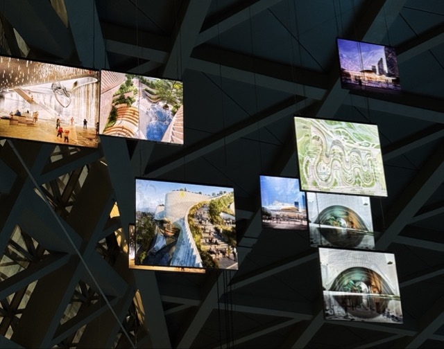 Multiple illuminated screens hanging from a ceiling structure, displaying images of architectural designs and landscapes.