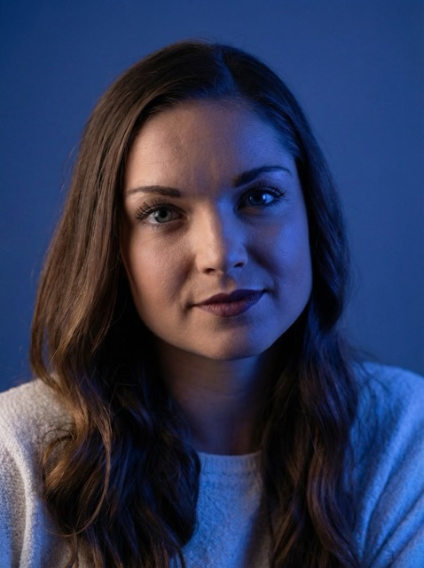 Portrait of a woman with long brown hair and a white sweater against a blue background.