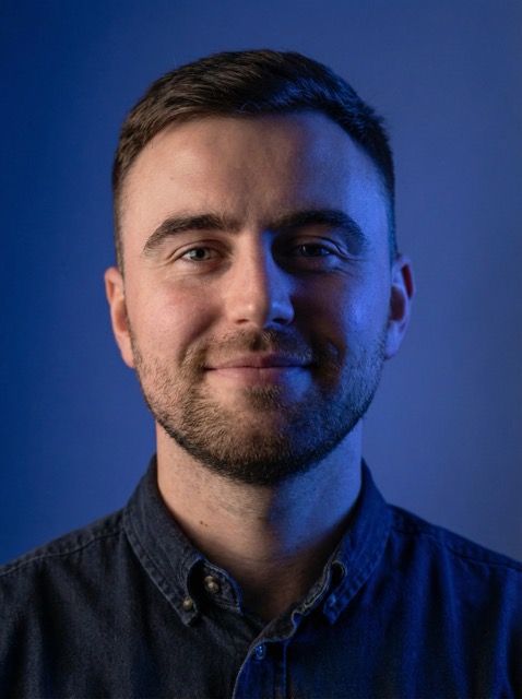 Smiling man with short brown hair and beard wearing a dark button-up shirt against a blue background.