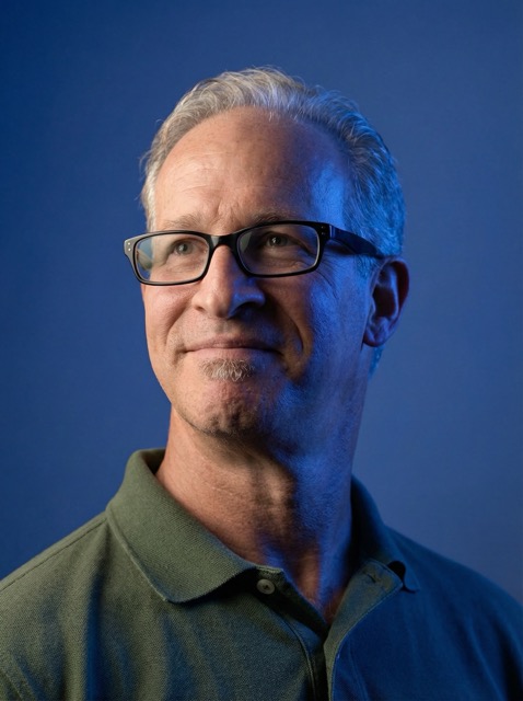 Middle-aged man with gray hair and glasses wearing a green polo shirt, looking slightly to the side against a blue background.