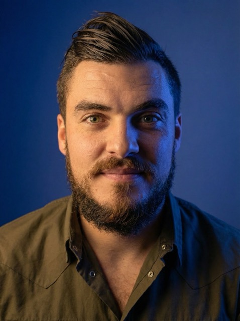 Portrait of a man with a beard and styled hair wearing an olive-green button-up shirt against a dark blue background.