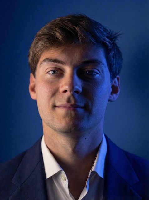 Portrait of a young man with light brown hair wearing a navy blue blazer and white shirt against a dark blue background.