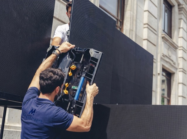 Two men assembling a large outdoor LED screen on a building facade.