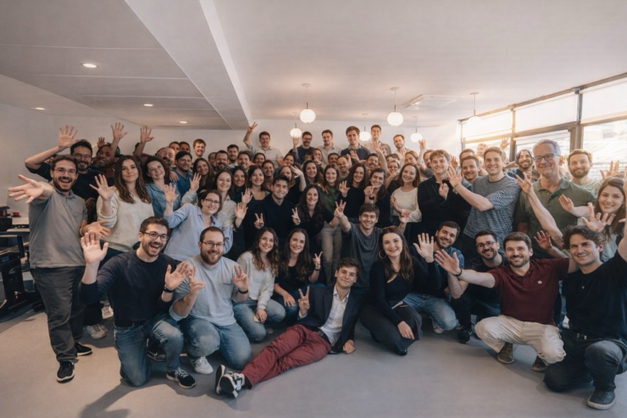 Large group of diverse people gathered in a bright office space smiling and waving at the camera.