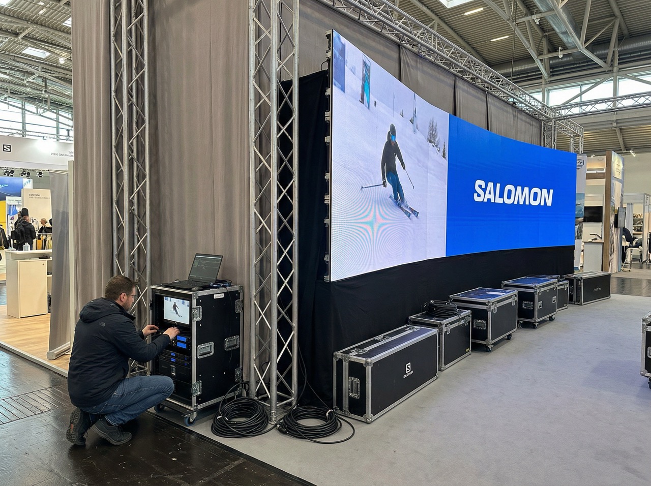 Man adjusting equipment near a large curved LED screen displaying a skier and the Salomon logo in an indoor exhibition space.