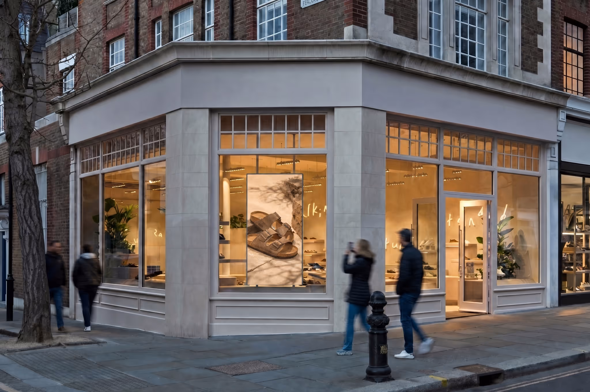 Corner view of a shoe store with large windows showing illuminated interior and a poster of brown sandals in the window.