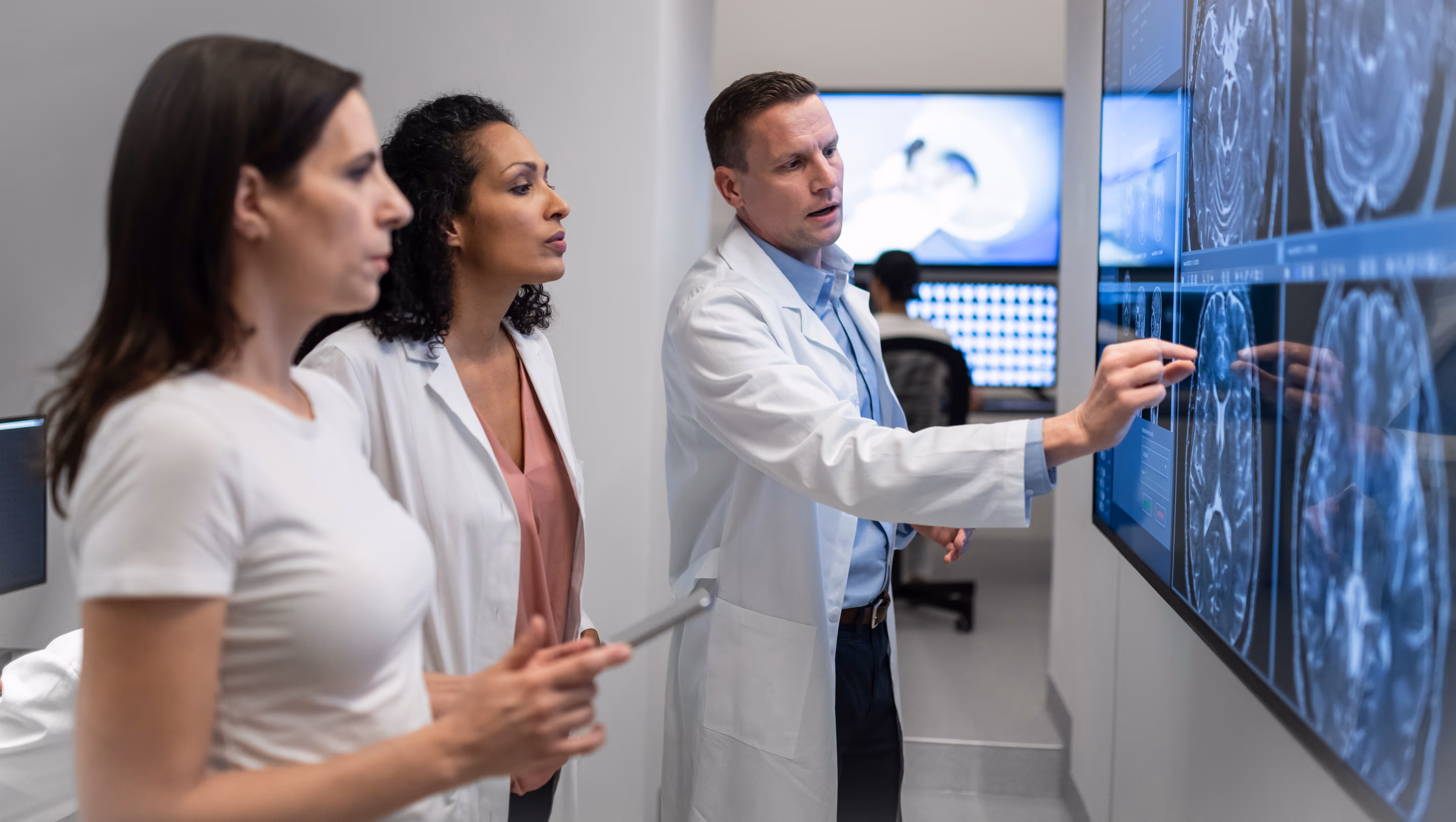 Two doctors, one man and one woman, point to medical imaging on the wall as a female patient watches.