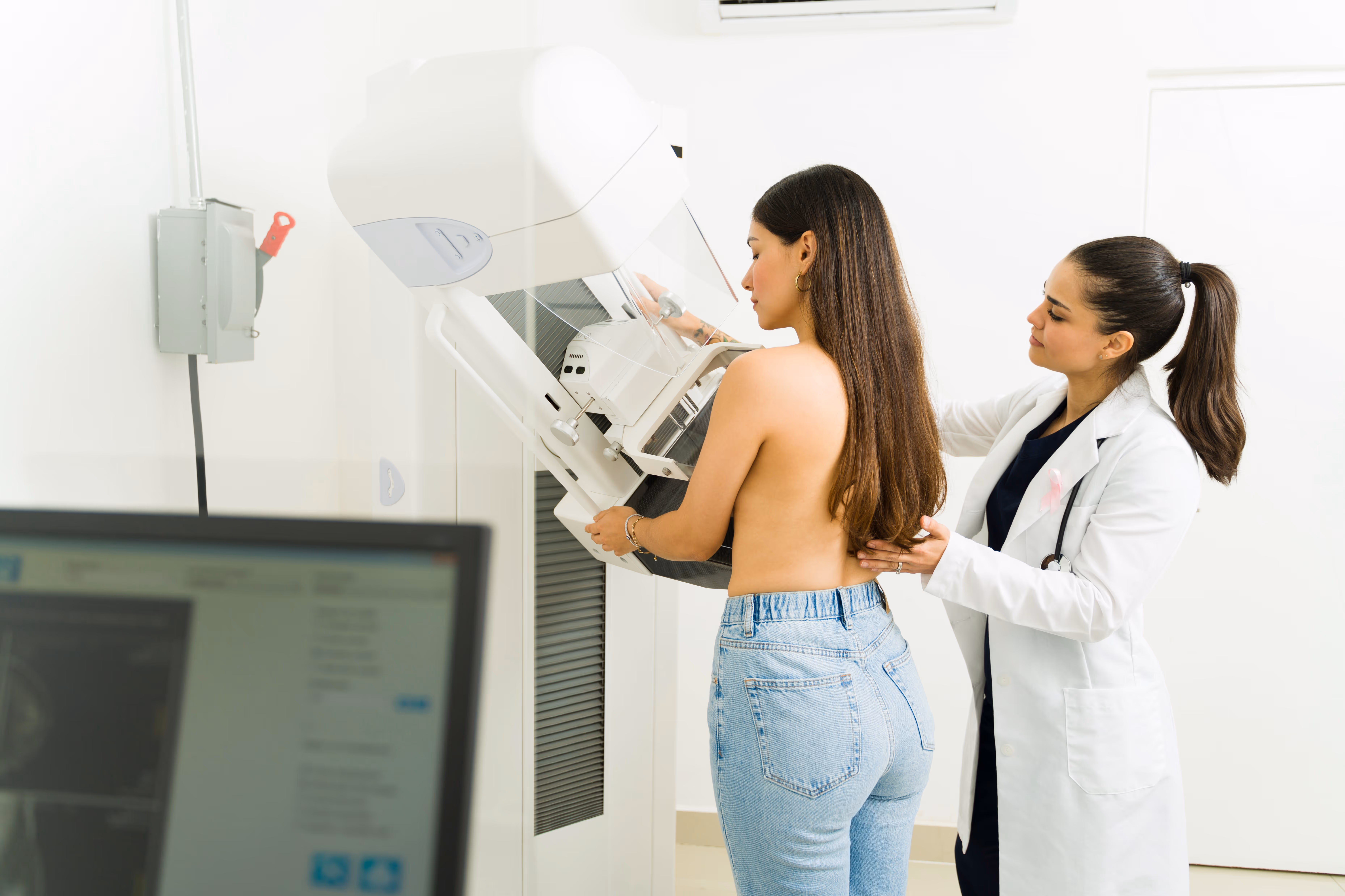 Dark haired woman getting a mammorgram while being helped by a female provider in lab coat.