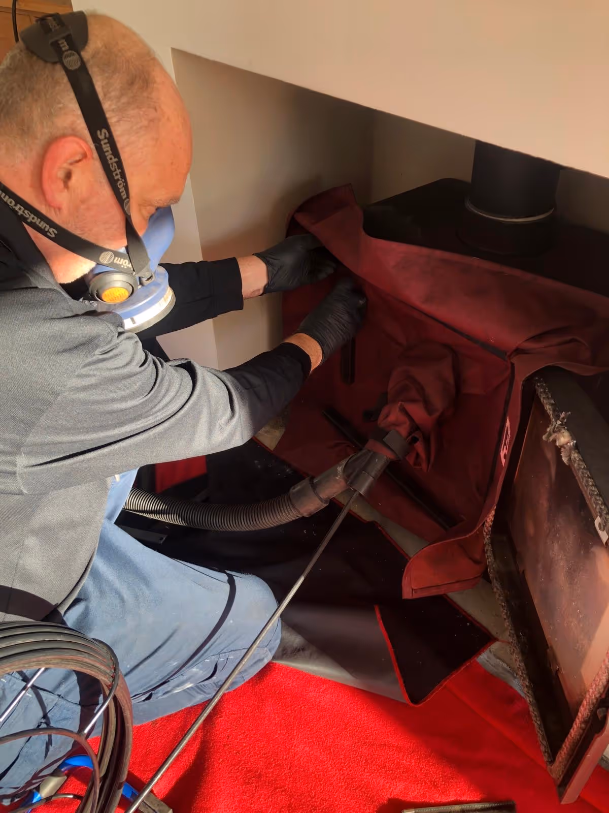 Person wearing a respirator mask and gloves cleaning or inspecting a fireplace with a hose and protective covers.