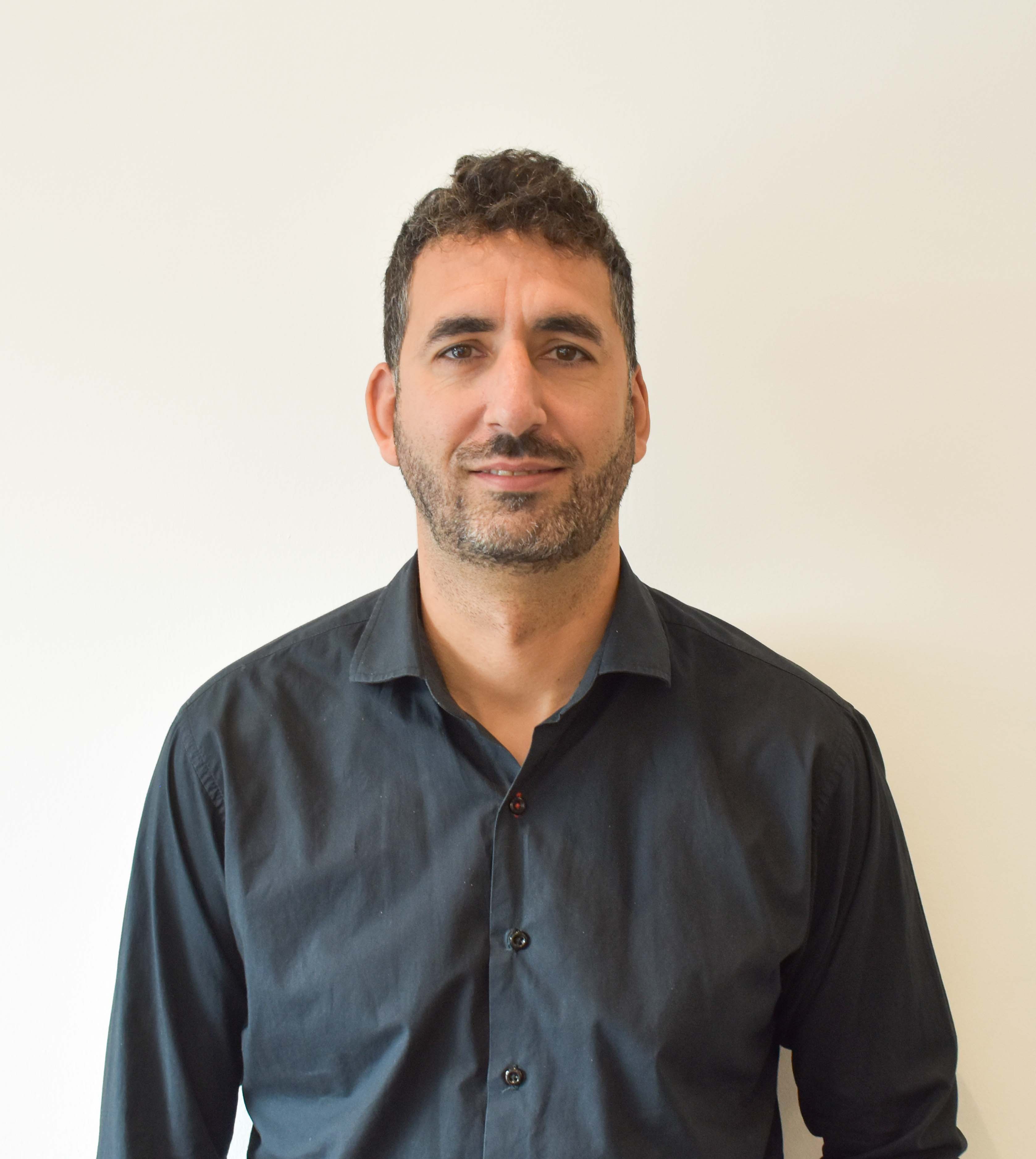 Hombre con barba y cabello rizado corto, viste camisa negra, frente a fondo blanco.