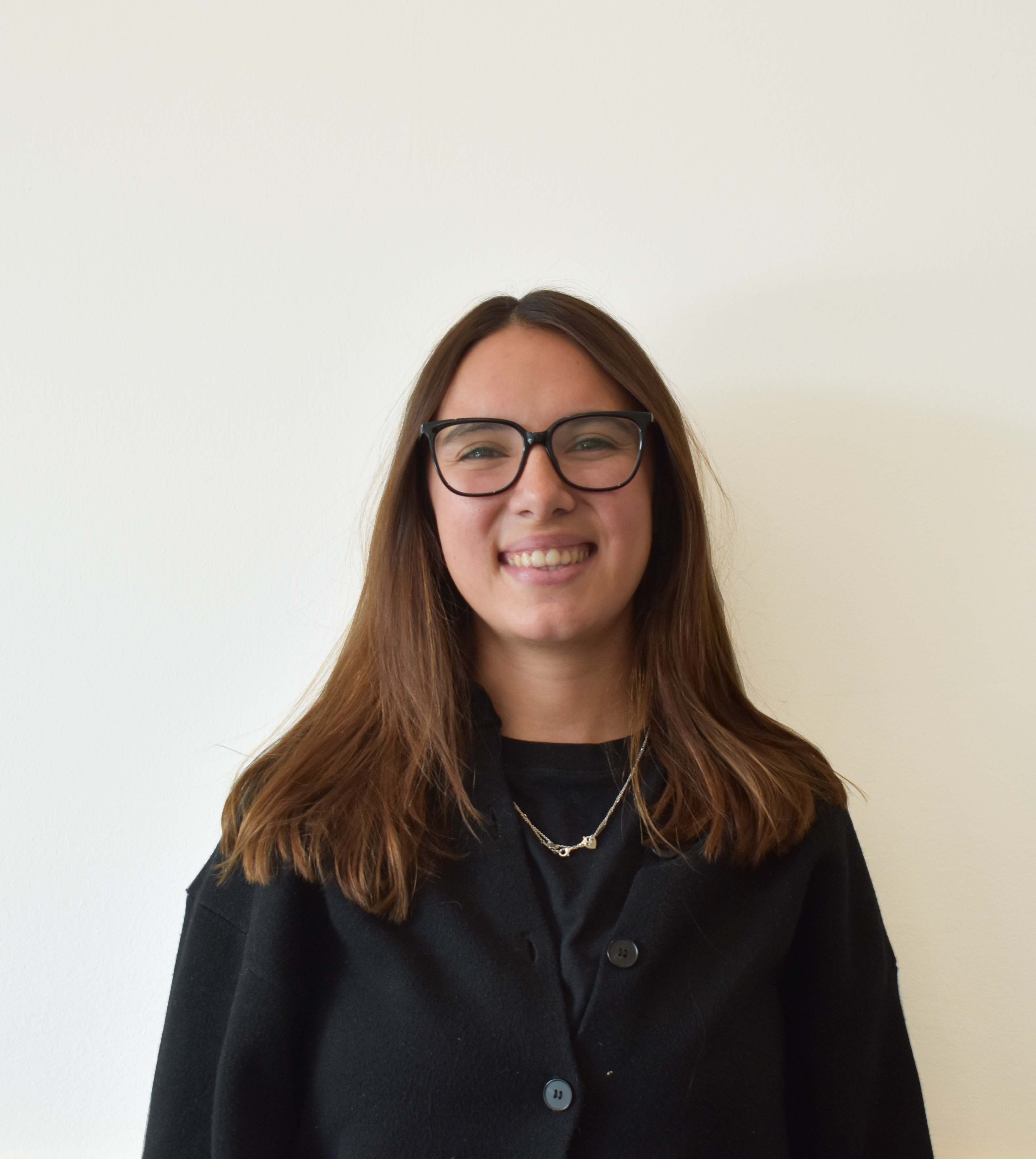 Mujer joven sonriente con cabello castaño, gafas negras y suéter negro frente a fondo claro.