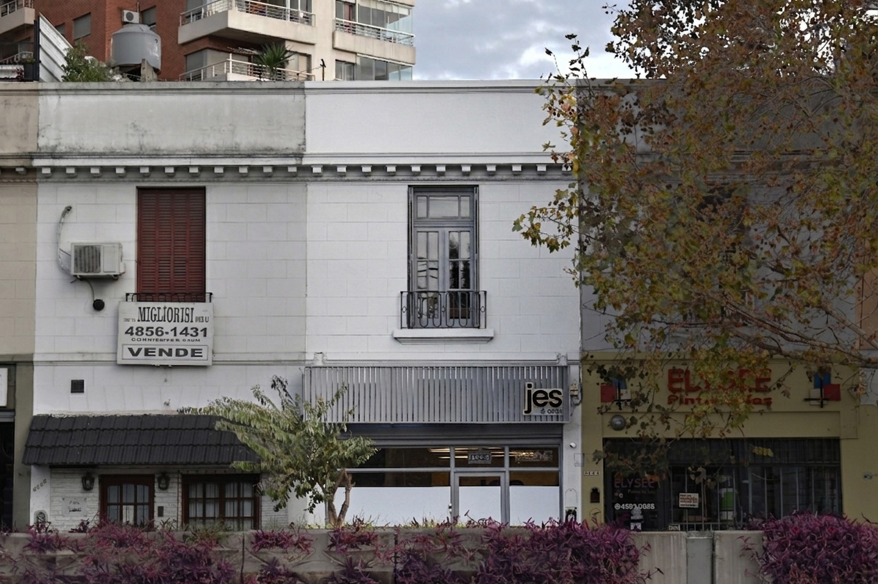 Fachada de edificio con cartel de venta, árbol y negocios al frente en un día nublado.