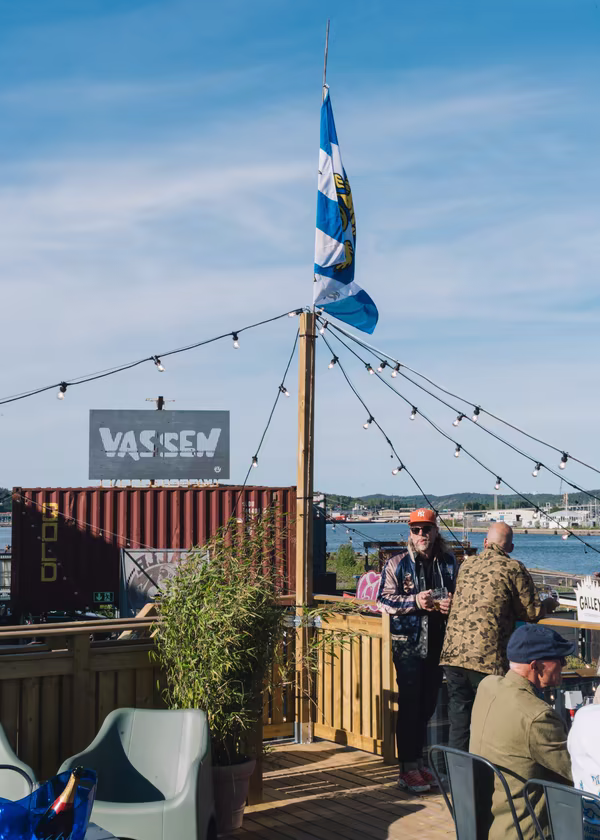 Outdoor waterfront seating area with people, string lights, a flag, and a red shipping container marked VASSEM.
