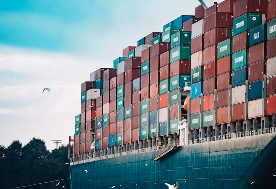 Large cargo ship loaded with stacked multicolored shipping containers on a clear day with birds flying nearby.