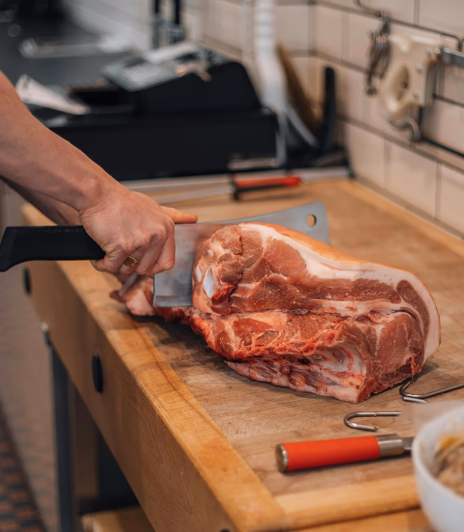 Person cutting a large raw pork shoulder on a wooden butcher block with a cleaver.