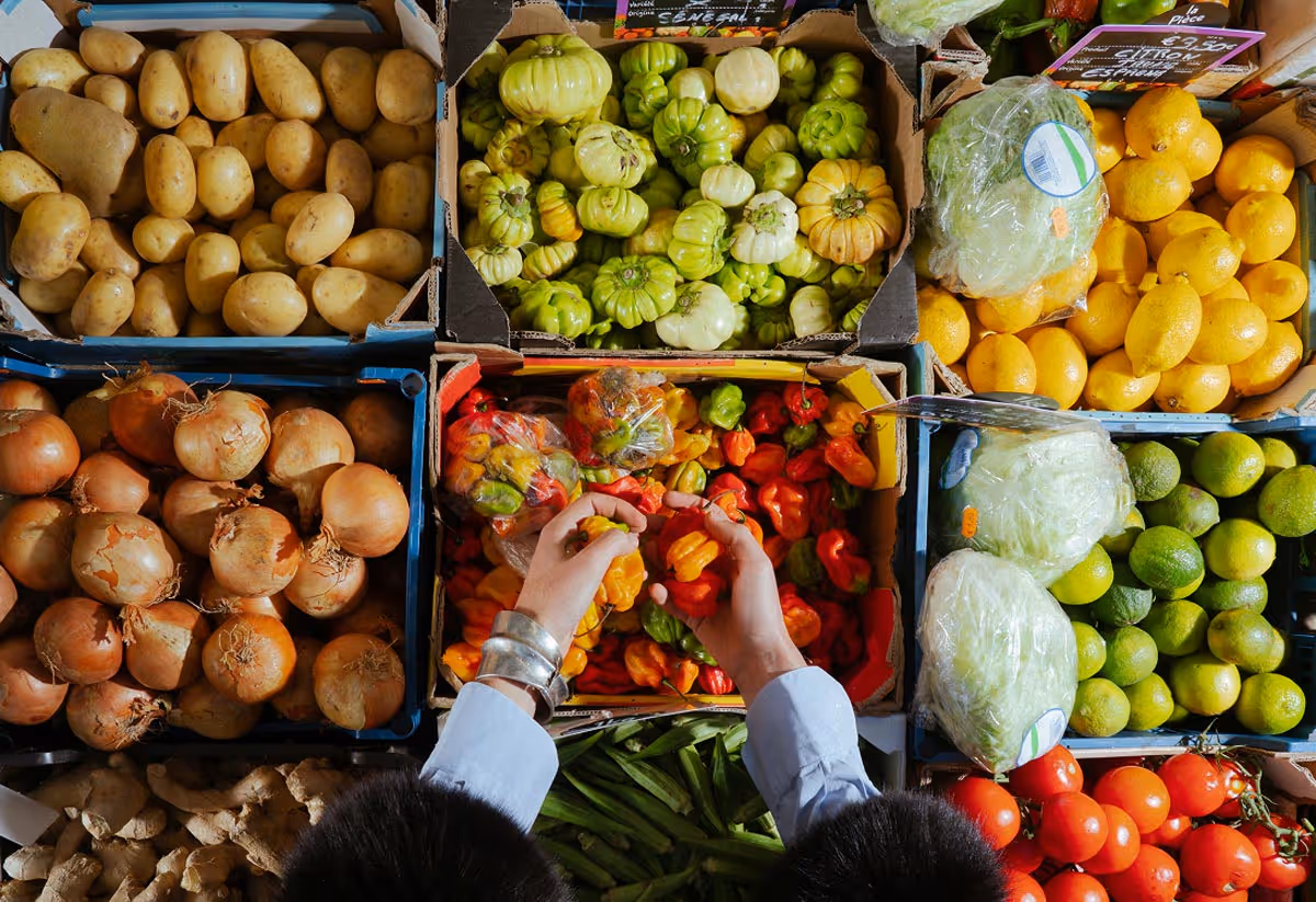 Person wearing silver bracelets selecting colorful bell peppers from a box surrounded by potatoes, onions, lemons, lettuce, limes, okra, and tomatoes at a market.