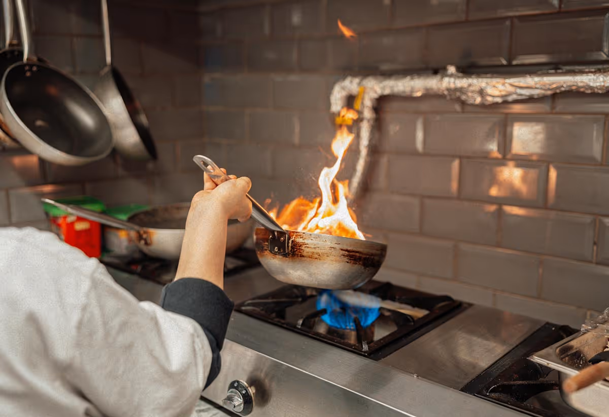 Chef’s hand holding a frying pan with flames rising over a gas stove in a kitchen.