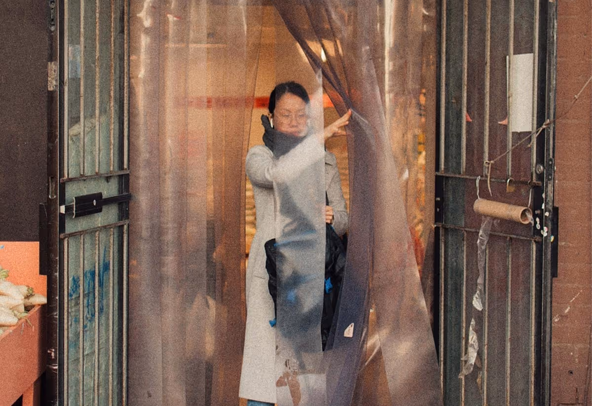Woman wearing glasses and a gray coat walking through a plastic strip curtain in a doorway.