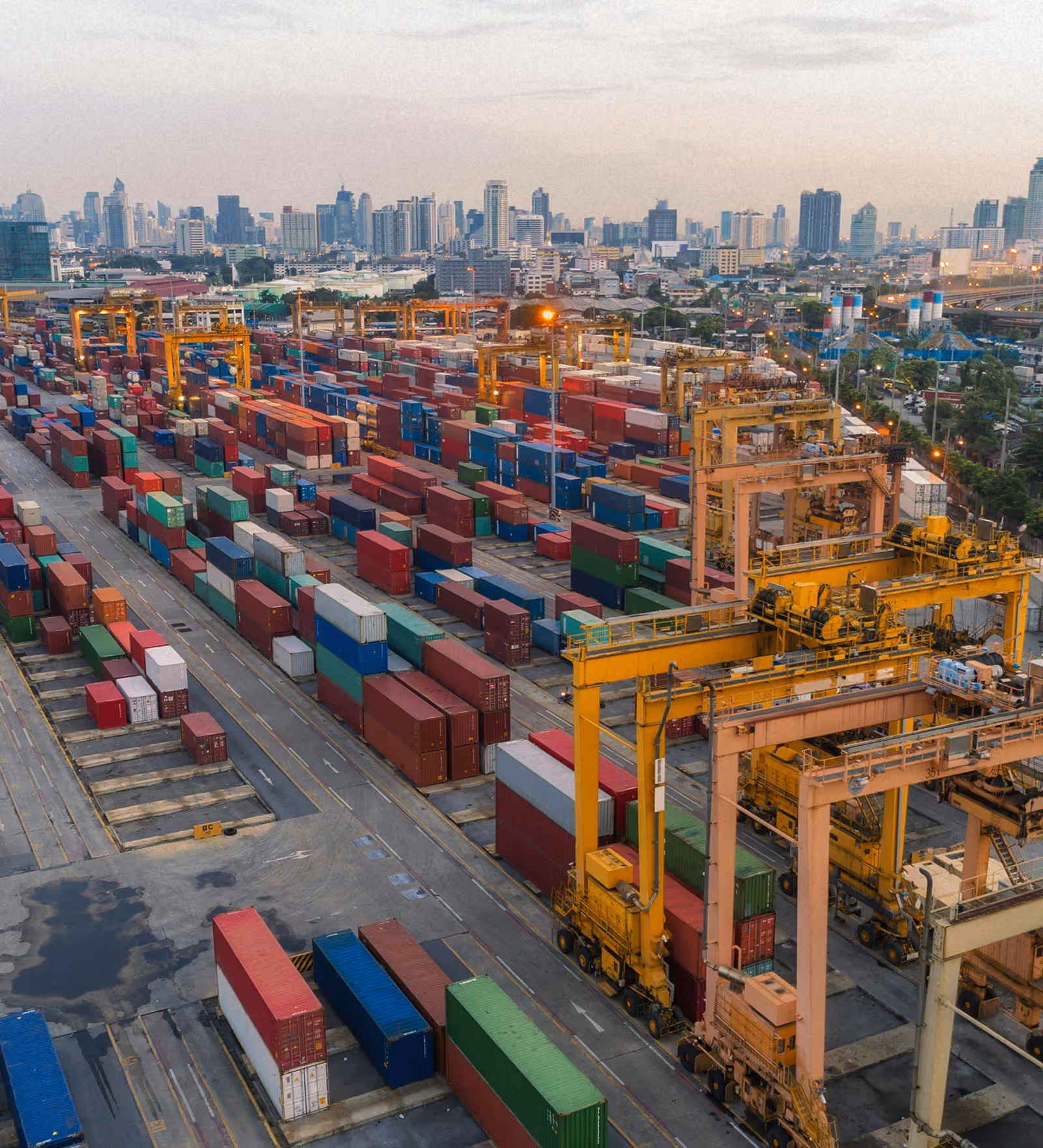 Aerial view of a busy shipping container yard with colorful containers and yellow cranes against a city skyline at dusk.