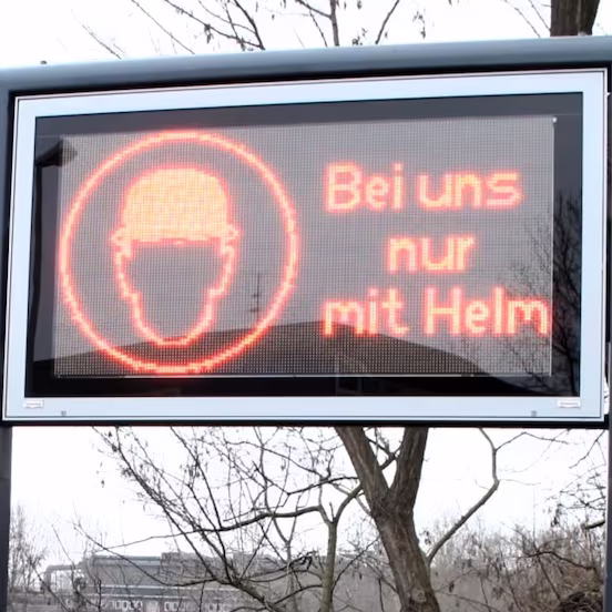 LED-Anzeigetafel auf einem Mast im Freien mit rotem Helm-Piktogramm und Text Bei uns nur mit Helm