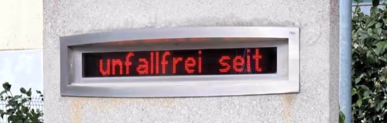Längliche LED-Laufschrift in Edelstahlrahmen an einer Betonwand, roter Text unfallfrei seit
