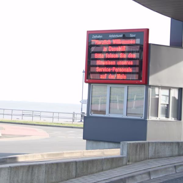 Red-framed LED display board above a harbour building with columns for destination port, time and lane