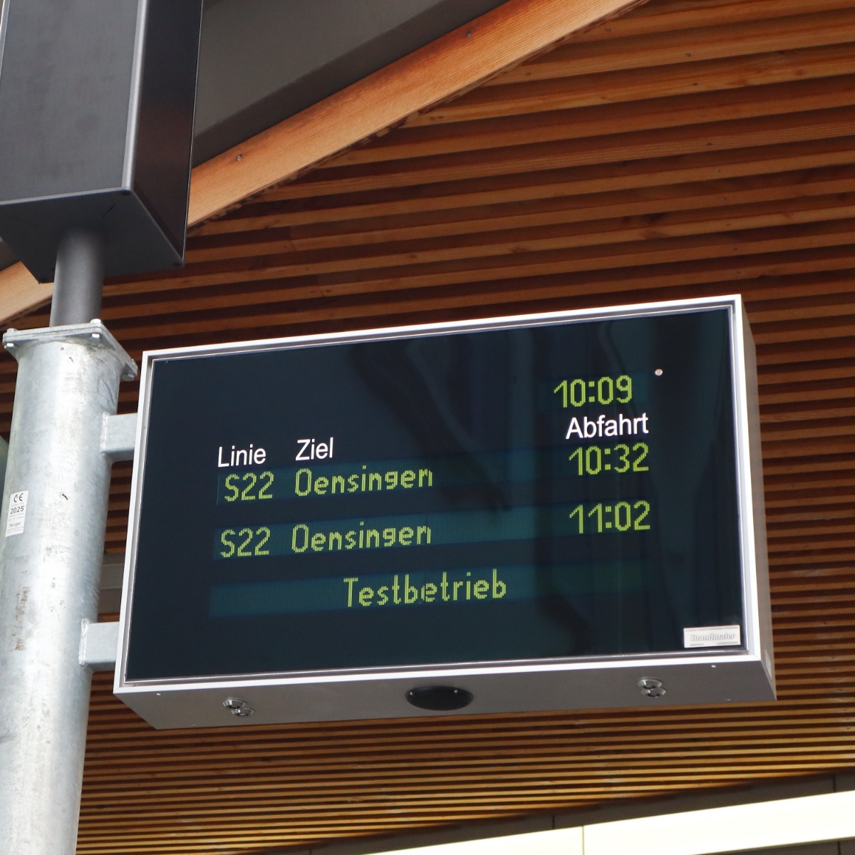 Pole-mounted display board in front of a wooden facade showing two departures and the note Testbetrieb