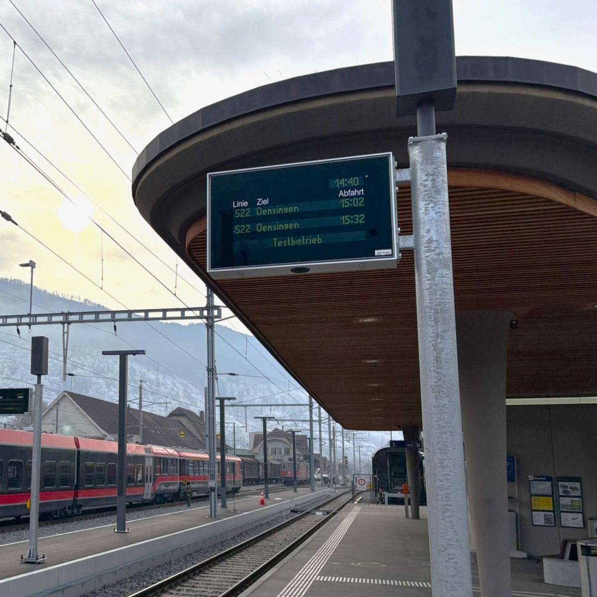 Platform with a curved canopy and a departure display mounted on it, a train and mountains behind