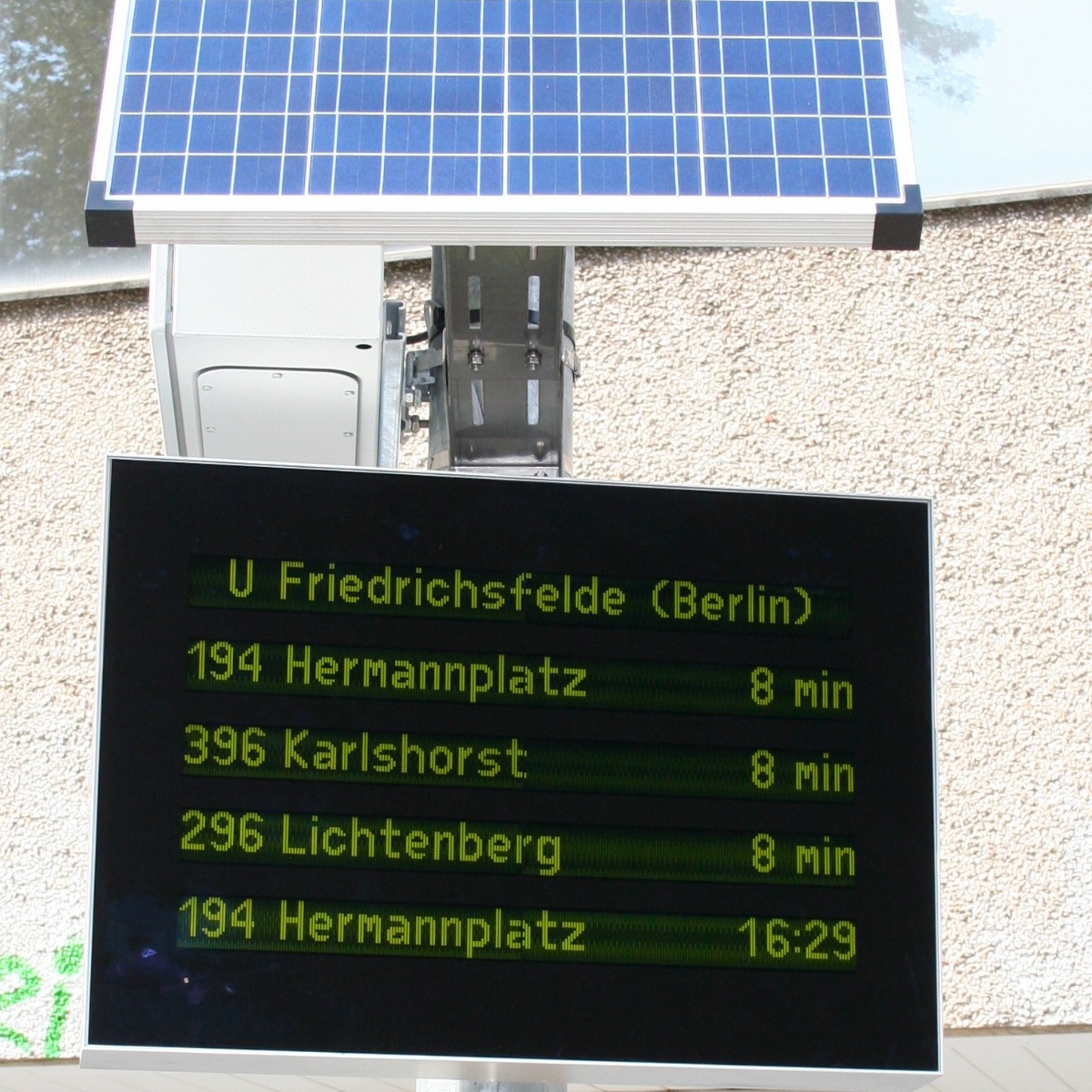 Display board with green text and a solar module mounted above it on a house wall