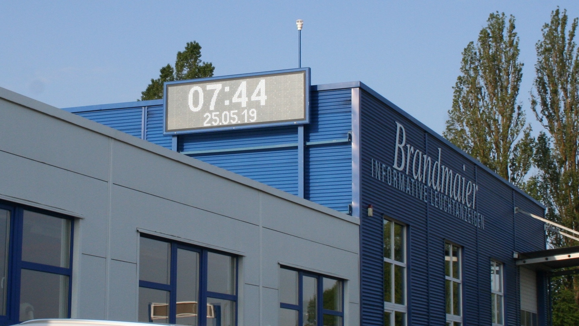 Digitale Uhrzeit- und Datumsanzeige auf dem Dach eines blauen Betriebsgebäudes mit Brandmaier-Schriftzug