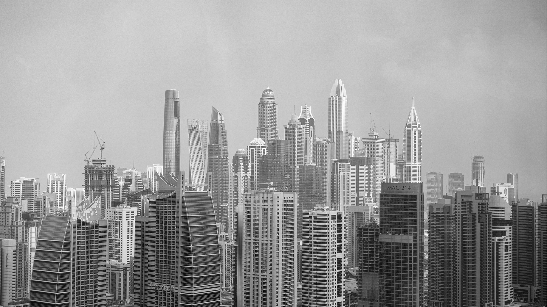 Black and white photo of a modern conference room with a wooden table, five leather office chairs, large TV, shelves, and windows showing city buildings.