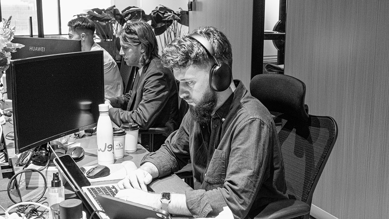 Man wearing headphones working on a laptop at a desk in an office with two other people working in the background.