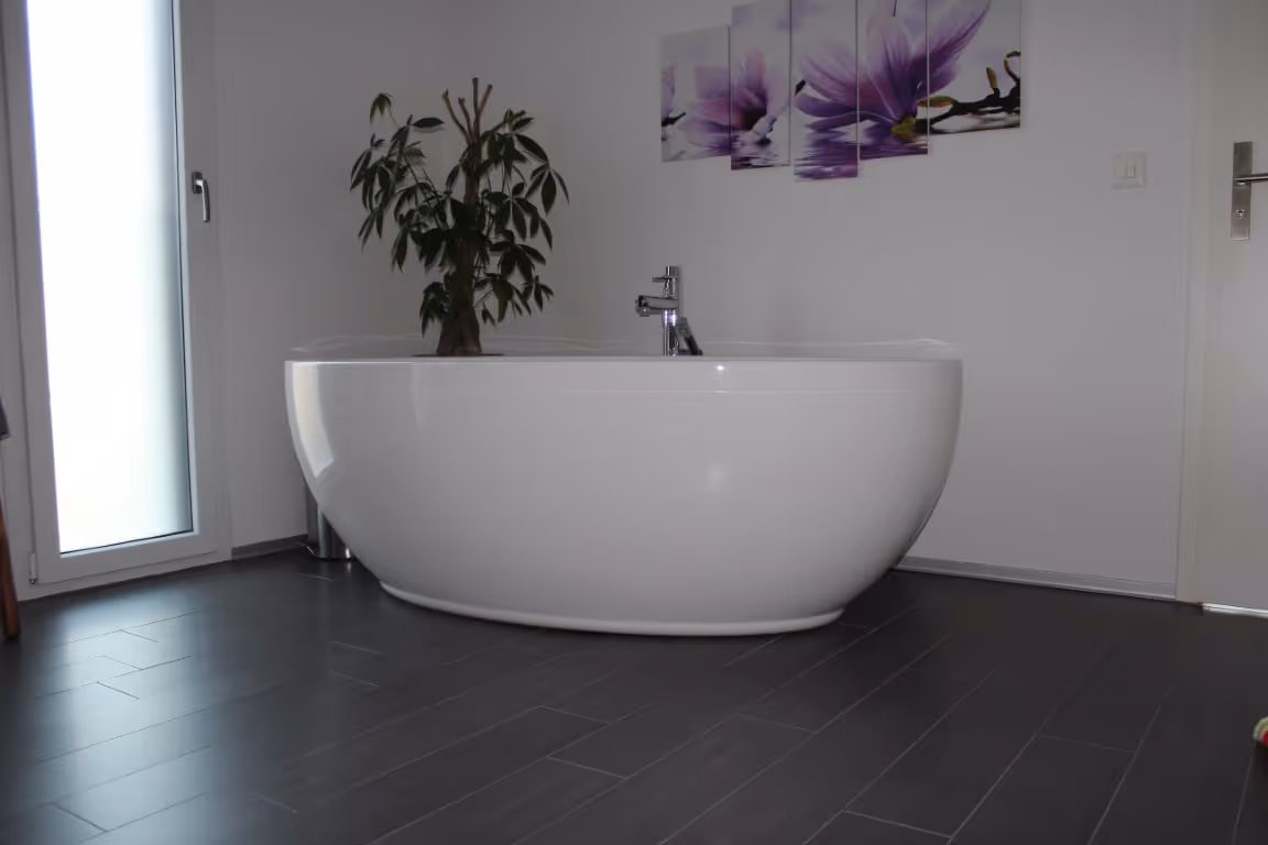 Moderne weiße freistehende Badewanne in einem Badezimmer mit dunklem Fliesenboden, einer Topfpflanze und violetter Blumenwandkunst.