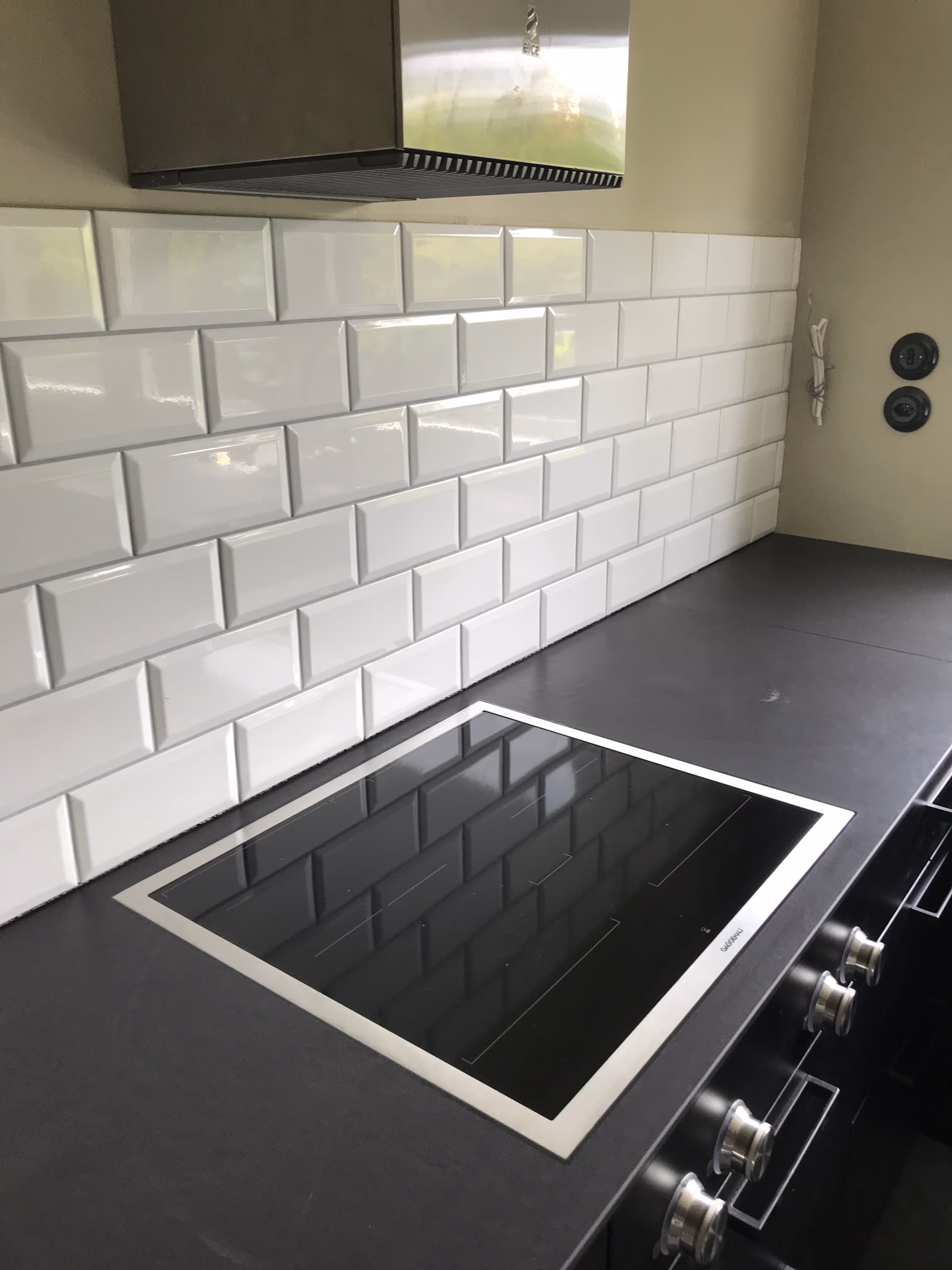 Moderne elektrische Kochplatte aus schwarzem Glas, installiert auf einer dunklen Arbeitsplatte mit weisser Fliesenrückwand und Edelstahl-Dunstabzugshaube darüber.