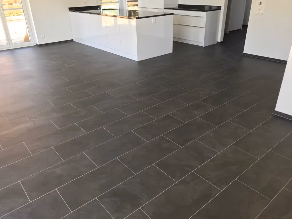 Moderne Küche mit weissen Schränken und schwarzer Arbeitsplatte, dunkelgrauem Fliesenboden und grossen Fenstern, die Tageslicht hereinlassen.