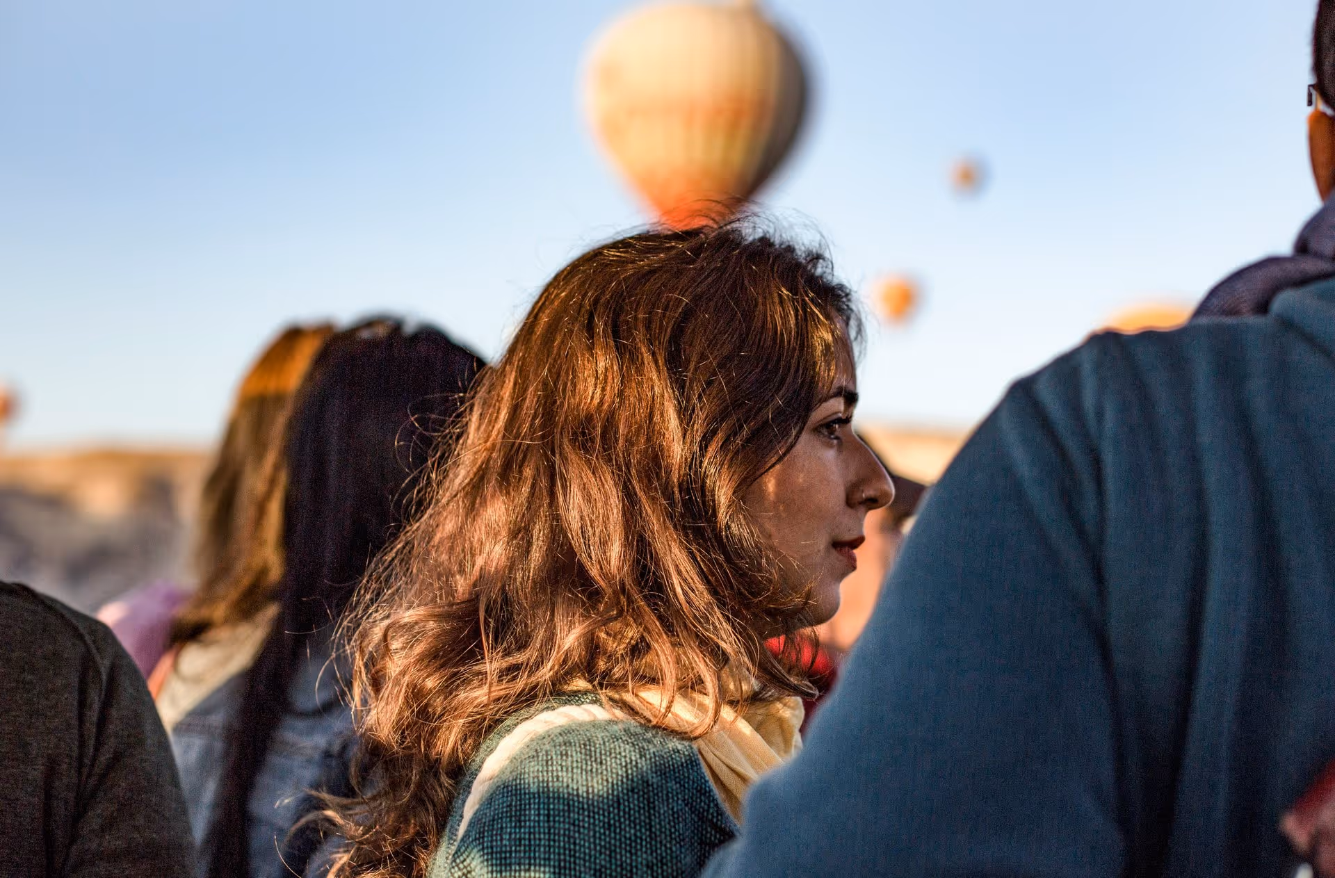 Profile of a woman with long brown hair, softly lit by the sun, among a crowd. Hot air balloons float in the clear blue sky above, conveying a sense of wonder.