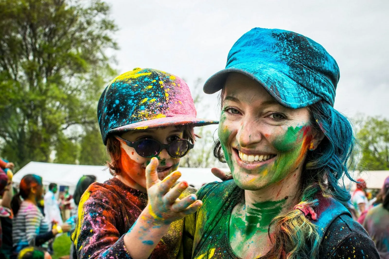 A woman and child, covered in vibrant colored powders, smile joyfully at a festival. They wear colorful caps and the background shows people and trees.