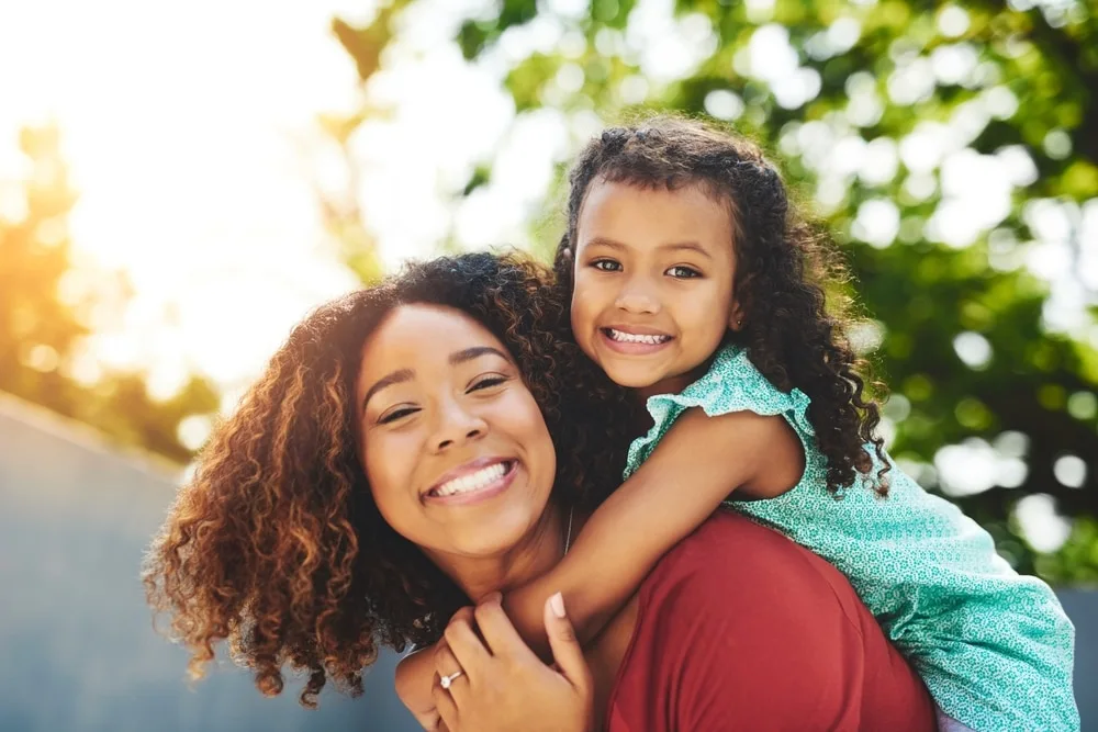 Nanny smiling with young girl on her back outdoors in natural light